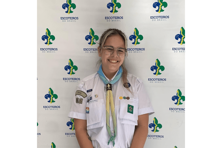 Imagem mostra uma menina com uniforme de escoteiro, camiseta branca e uma fita colorida em seu pescoço