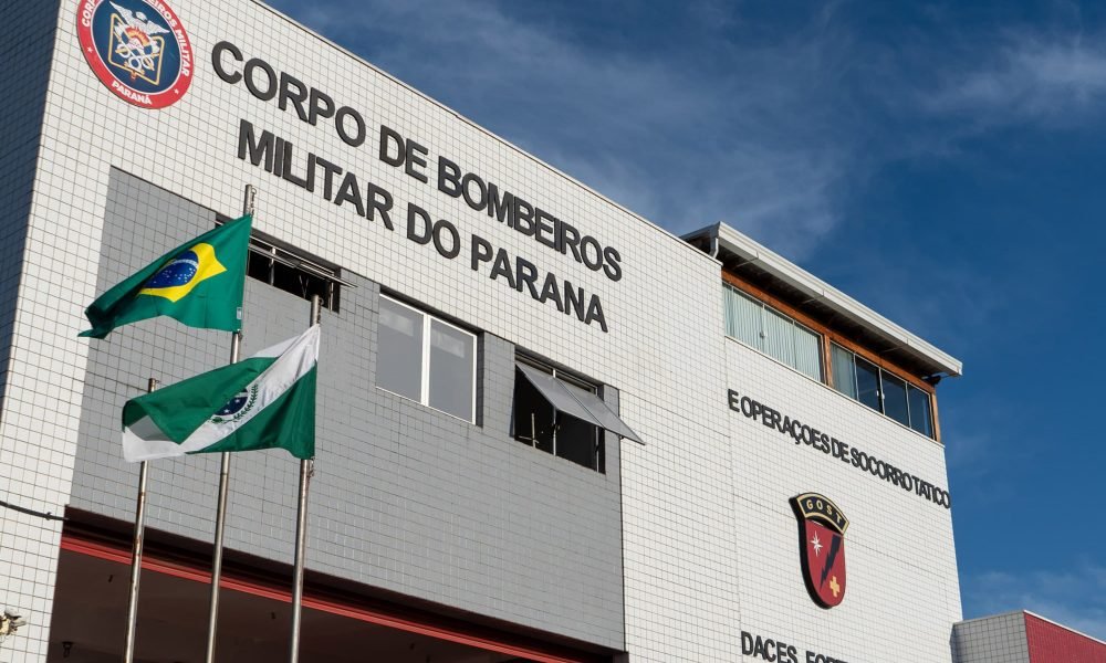 Imagem mostra a fachada do prédio do corpo de bombeiros com duas bandeiras hasteadas na frente, sendo elas do Paraná e do Brasil