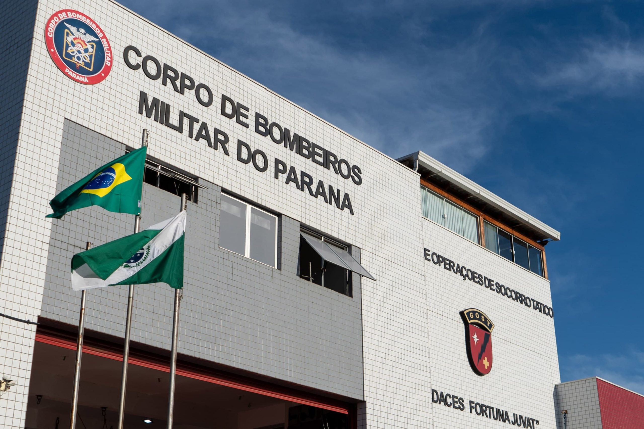 Imagem mostra a fachada do prédio do corpo de bombeiros com duas bandeiras hasteadas na frente, sendo elas do Paraná e do Brasil