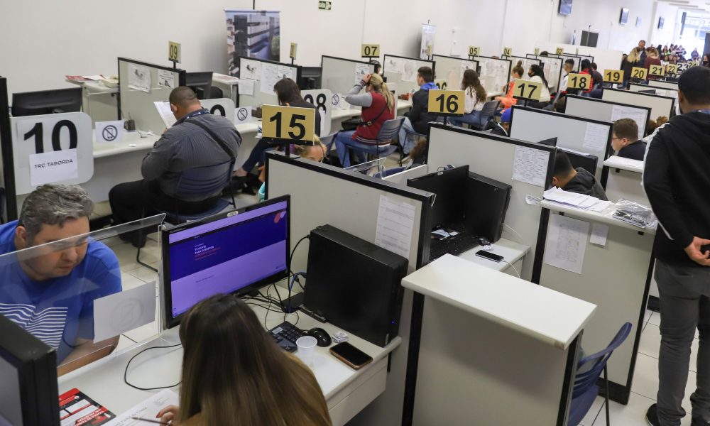 Imagem mostra uma sala branca com diversas mesas e cadeiras com pessoas atendendo outras pessoas