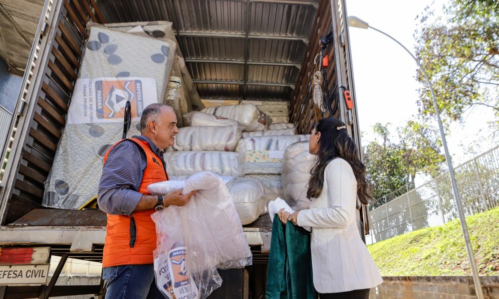 Tenente-coronel da defesa civil, usando um colete alaranjado e a primeira-dama do Paraná utilizando um casaco branco, em frente a um caminhão cheio de donativos pronto para partir ao estado do Rio Grade do Sul.