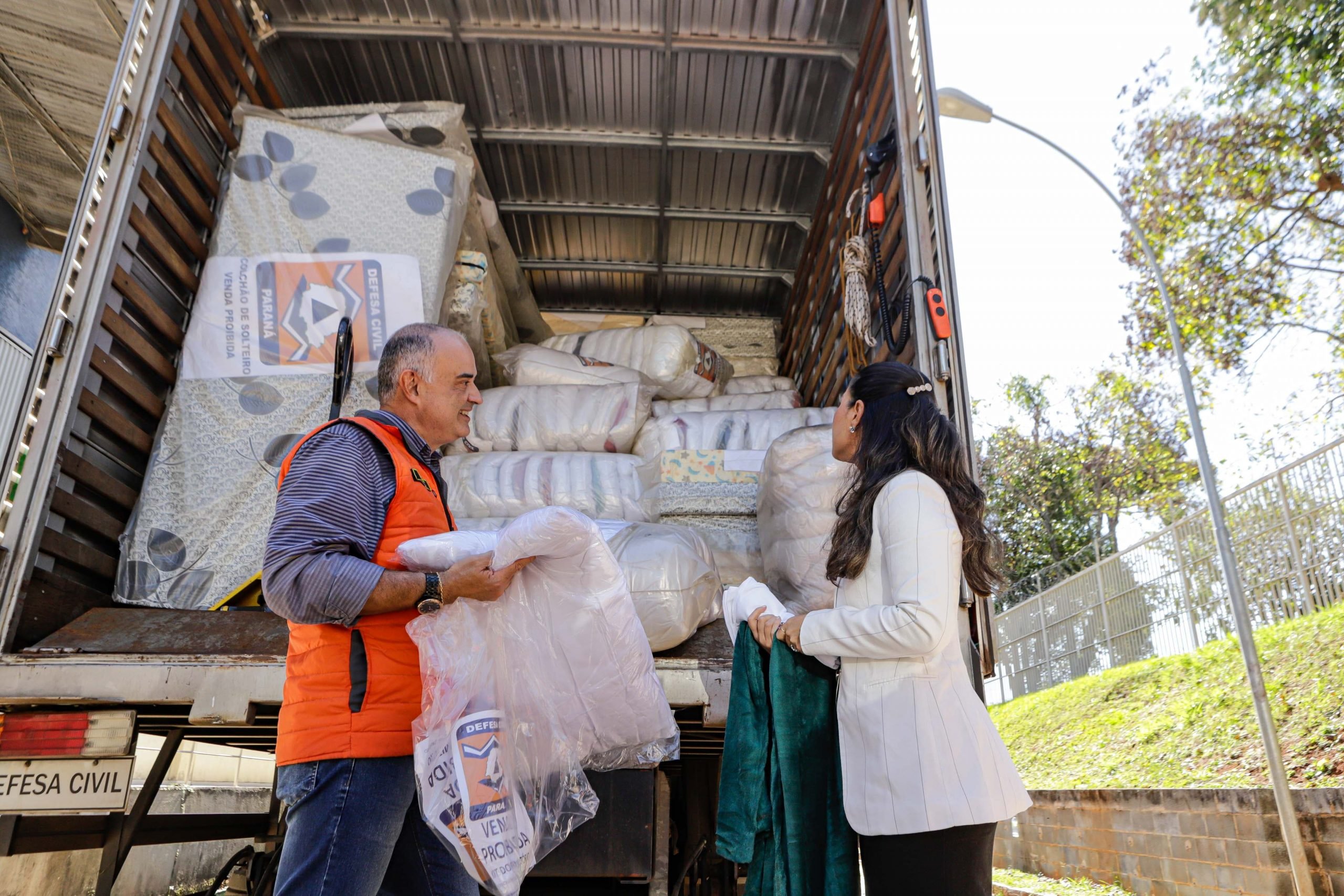 Tenente-coronel da defesa civil, usando um colete alaranjado e a primeira-dama do Paraná utilizando um casaco branco, em frente a um caminhão cheio de donativos pronto para partir ao estado do Rio Grade do Sul.