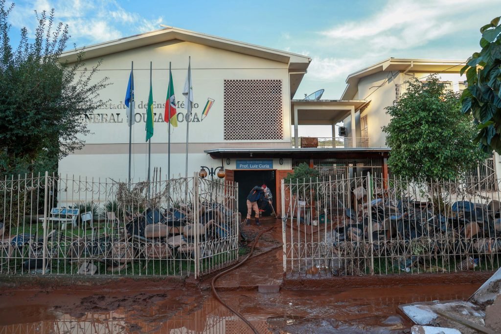 Imagem mostra a fachada de uma escola localizada no Rio Grande do Sul, onde há duas pessoas realizando a limpeza da escola.