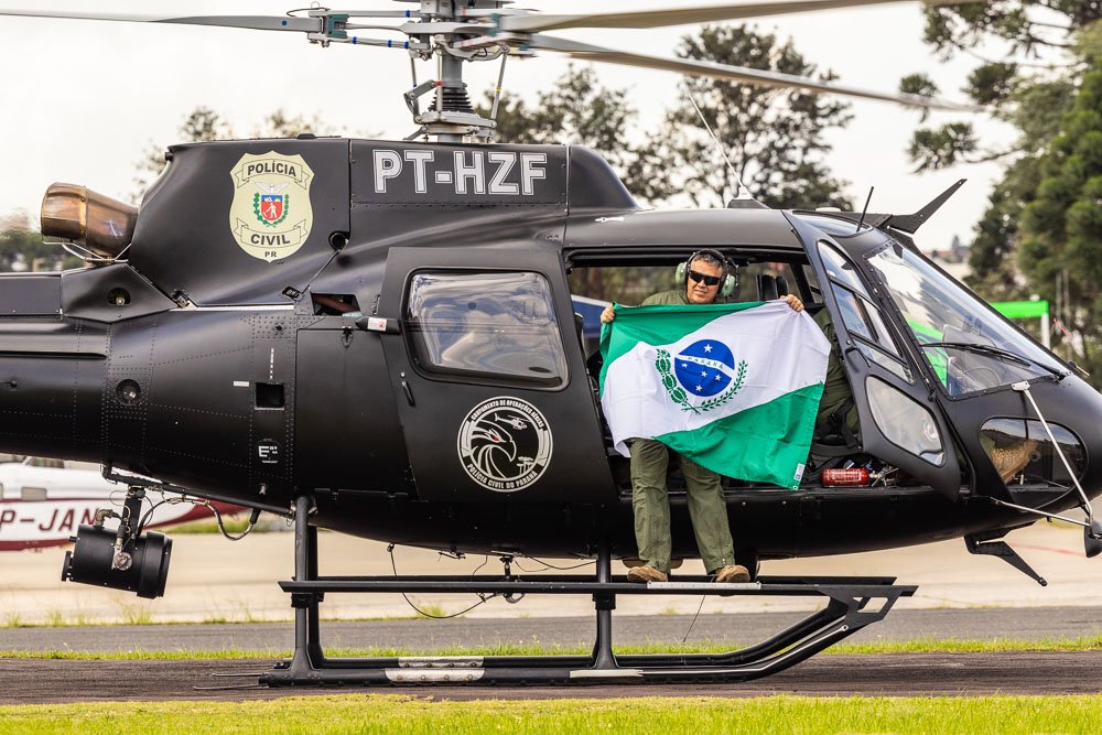 Helicóptero preto da PCPR com um homem segurando a bandeira do Paraná e sorrido