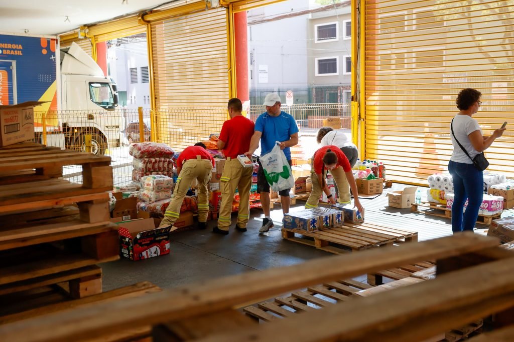 Corpo de Bombeiros recebendo doações, vestindo camisetas vermelhas e voluntários auxiliando na organização das doações.