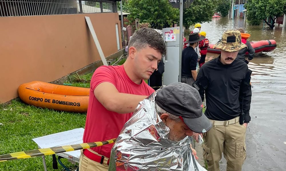 Bombeiro vestindo camisa vermelha cobrindo uma pessoa resgatada das enchentes com uma manta prata