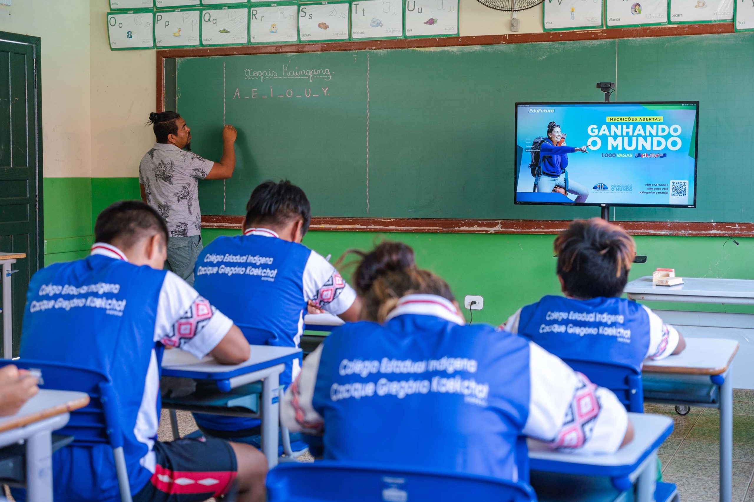 Imagem mostra parte de uma sala de aula de uma escola indígena, com o professor escrevendo no quadro. Mostrando que a taxa de analfabetismo tem diminuido