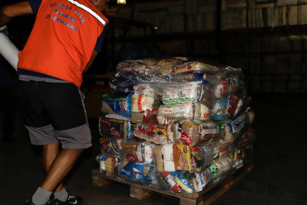 Trabalhador usando colete alaranjado escrito defesa civil colocando os donativos em um caminhão.