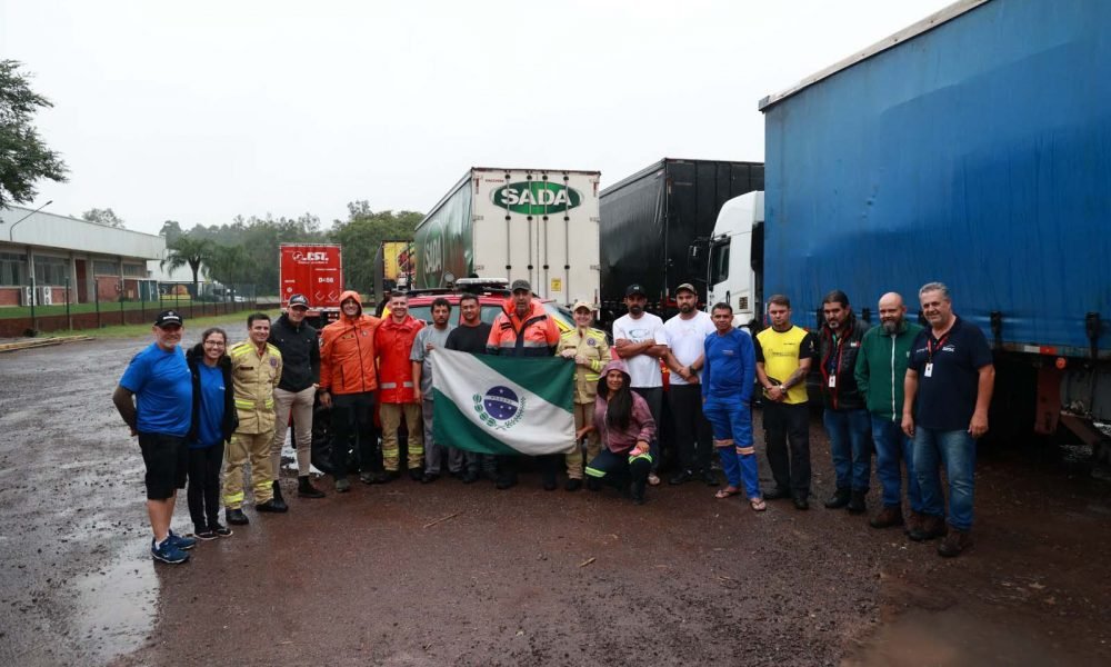 Imagem mostra diversas pessoas uma ao lado da outra, com a bandeira do Paraná na frente. São os voluntários que estão auxiliando na rede de ajuda humanitária