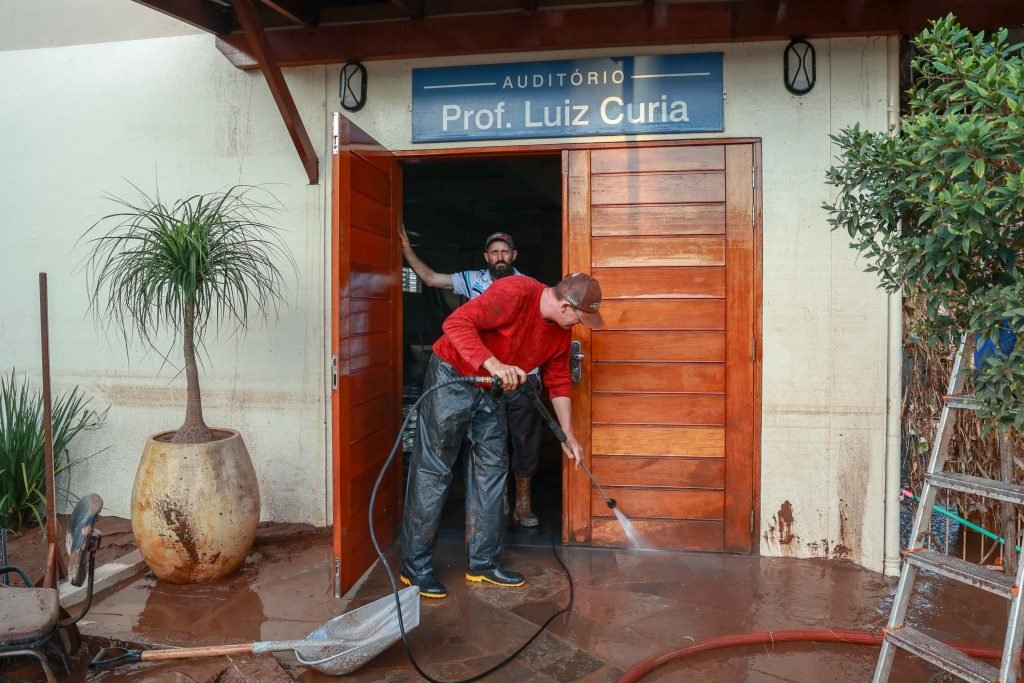 Imagem mostra a fachada de uma escola localizada no Rio Grande do Sul, onde há duas pessoas realizando a limpeza da escola.