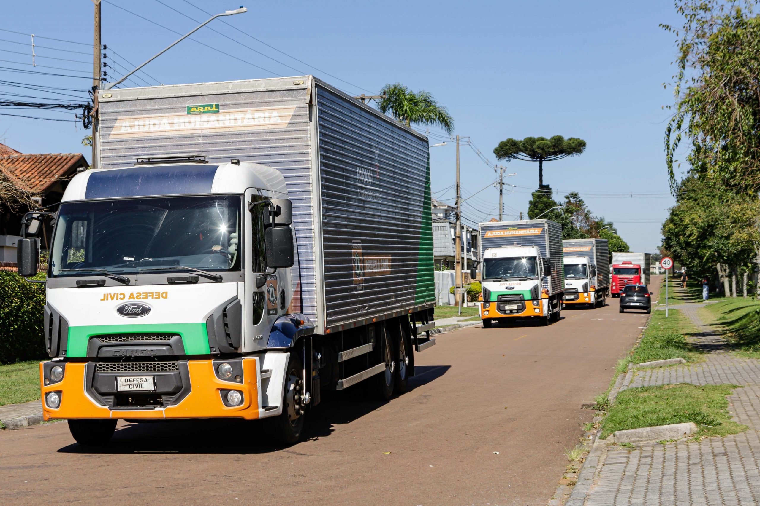 Caminhão com a frente branca, verde e amarela em forma de faixa, com a caçamba na cor prata e diversos outros caminhões atras prontos para levar os donativos ao Rio Grande do Sul