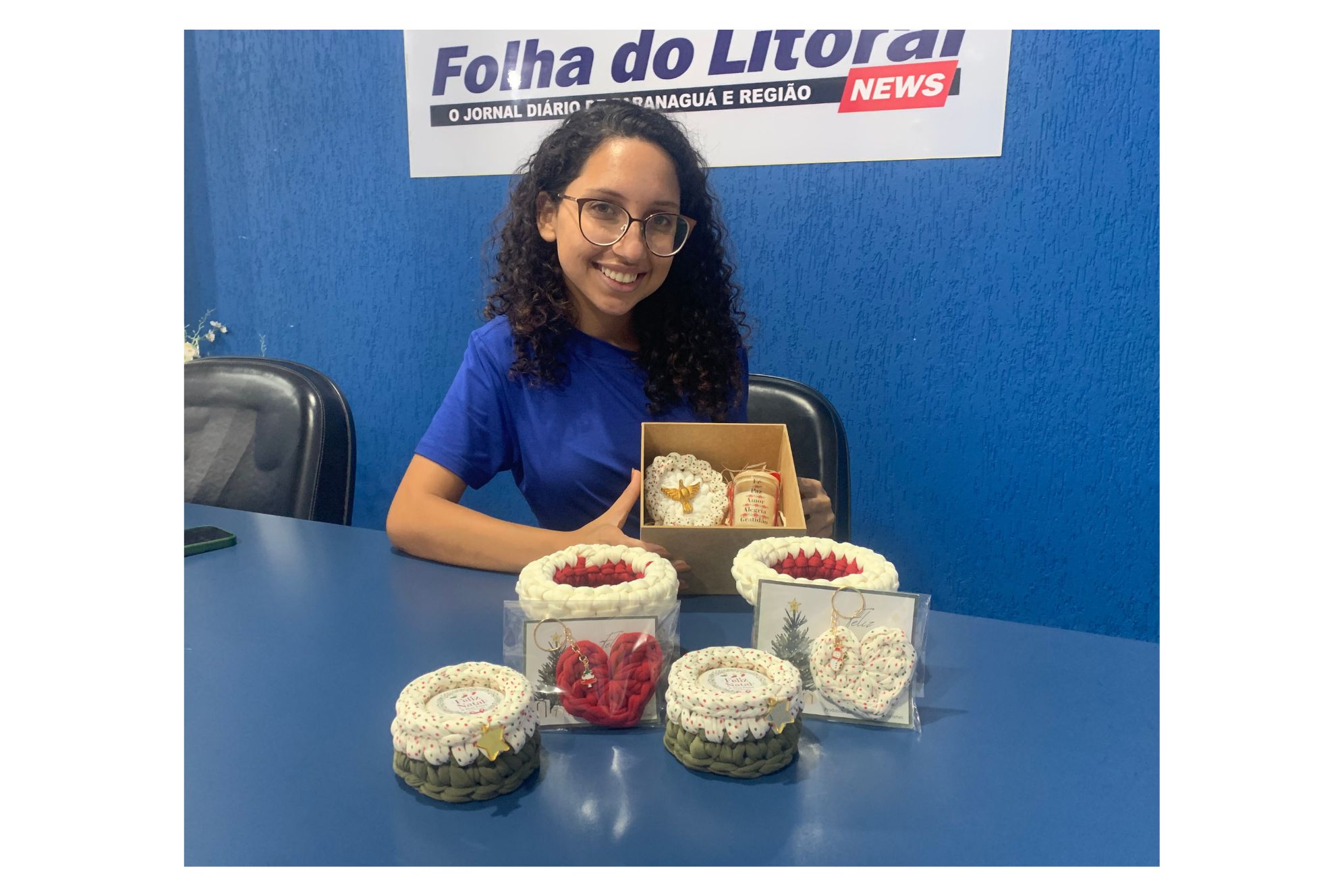 Uma jovem sorridente com cabelos cacheados usa óculos e segura uma caixa enquanto se senta em uma mesa com doces decorativos. Os doces estão dispostos em frascos e a jovem parece feliz em apresentar suas criações. Esta imagem destaca a habilidade artesanal em confeitaria e o prazer em compartilhar essas delícias. Artesã usa parte do seu lucro de vendas natalinas em ações voluntárias