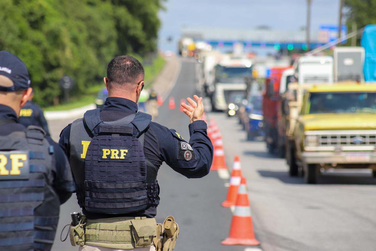 Agentes da PRF trabalham em uma rodovia, orientando caminhões em meio a sinalização de trânsito. Imagem destaca a importância da segurança nas estradas. Paraná recebe abertura nacional de operação para reduzir acidentes nas estradas