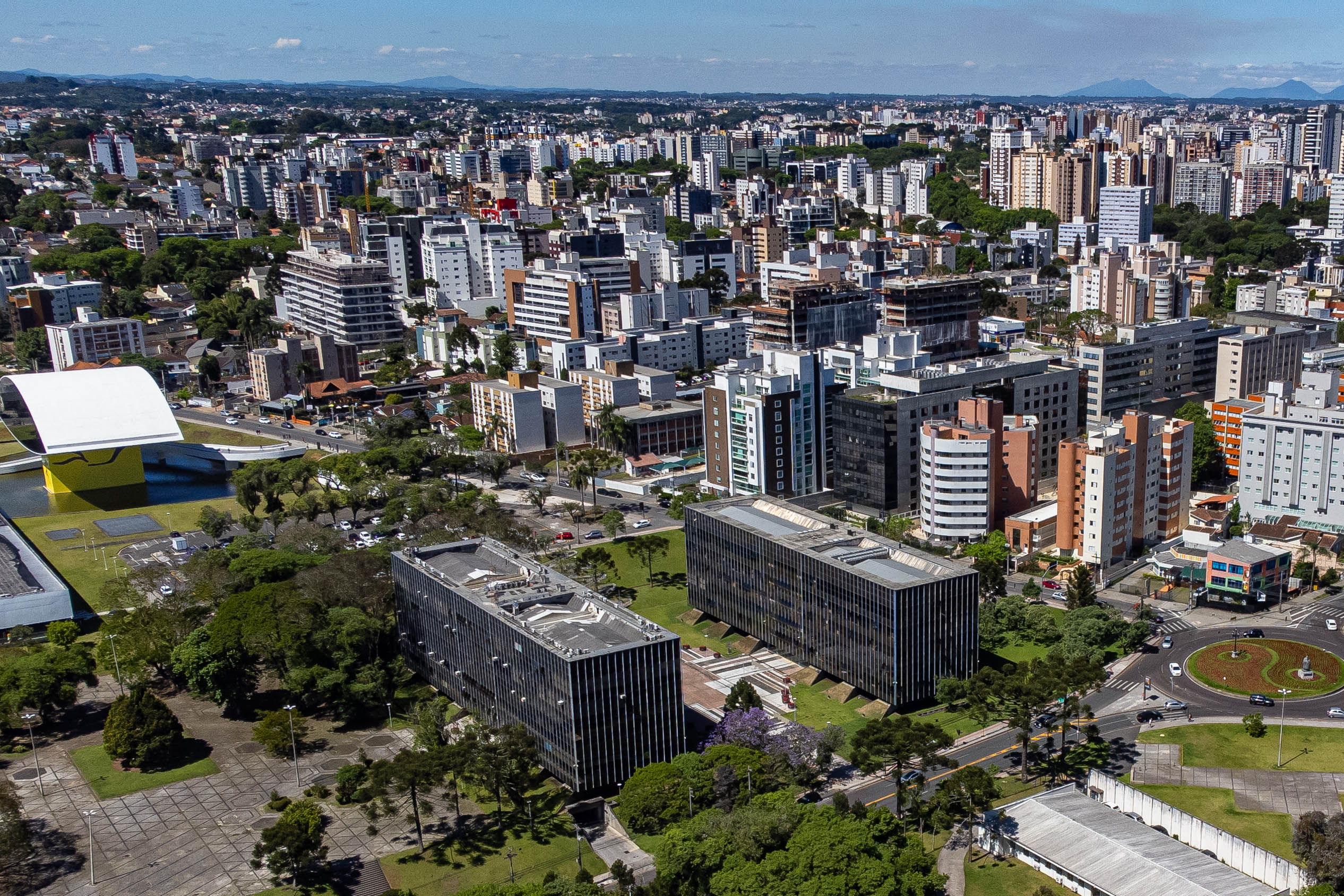 Vista aérea da cidade mostrando prédios, áreas verdes e a modernidade urbana. A imagem destaca a arquitetura contemporânea e urbanismo planejado.