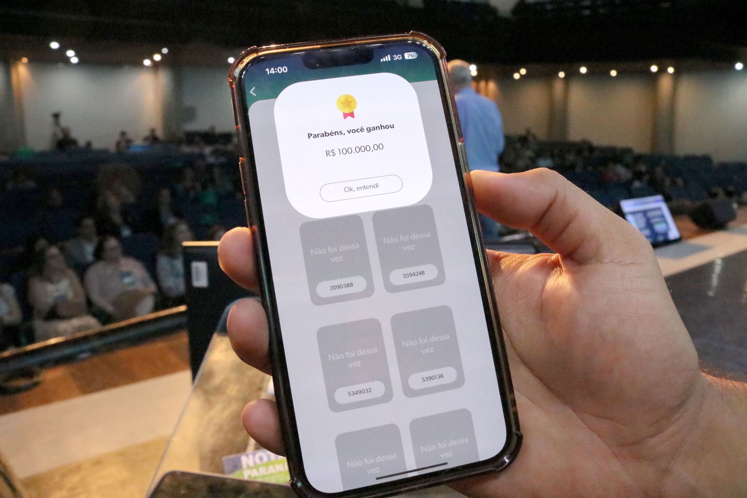 Imagem de um smartphone exibindo uma mensagem de vitória com o valor de R$ 100.000,00 na tela, em um evento ao vivo. A audiência ao fundo está atenta. R$ 133,3 milhões do Nota Paraná não foram resgatados pelos consumidores em 2024