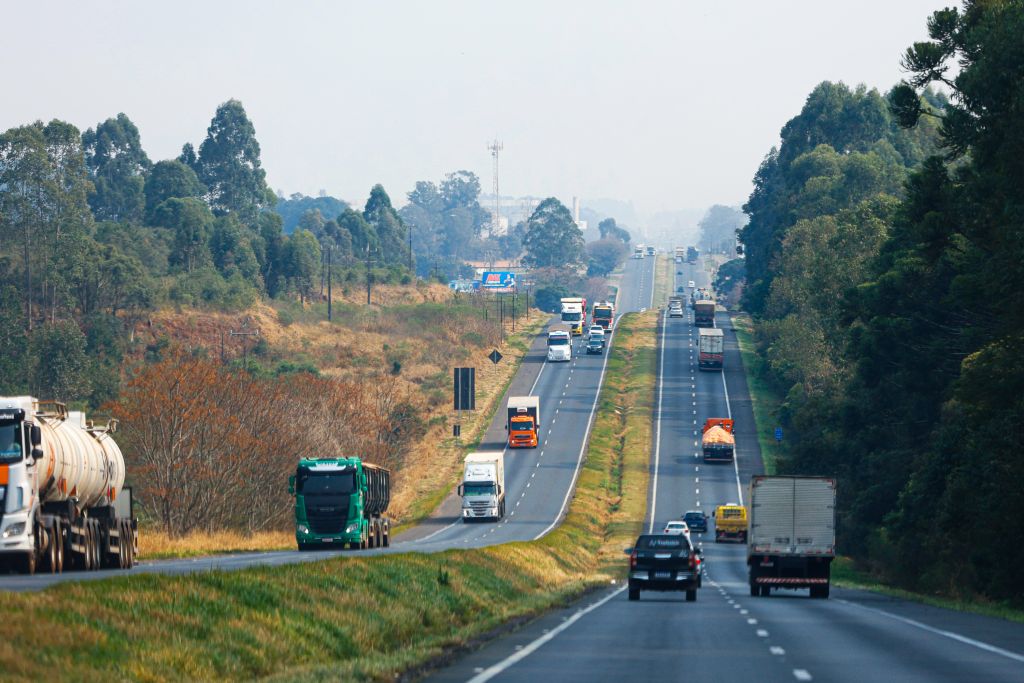 Rodovia movimentada com caminhões e veículos em uma bela paisagem verde, mostrando a importância do transporte rodoviário. Paraná recebe abertura nacional de operação para reduzir acidentes nas estradas
