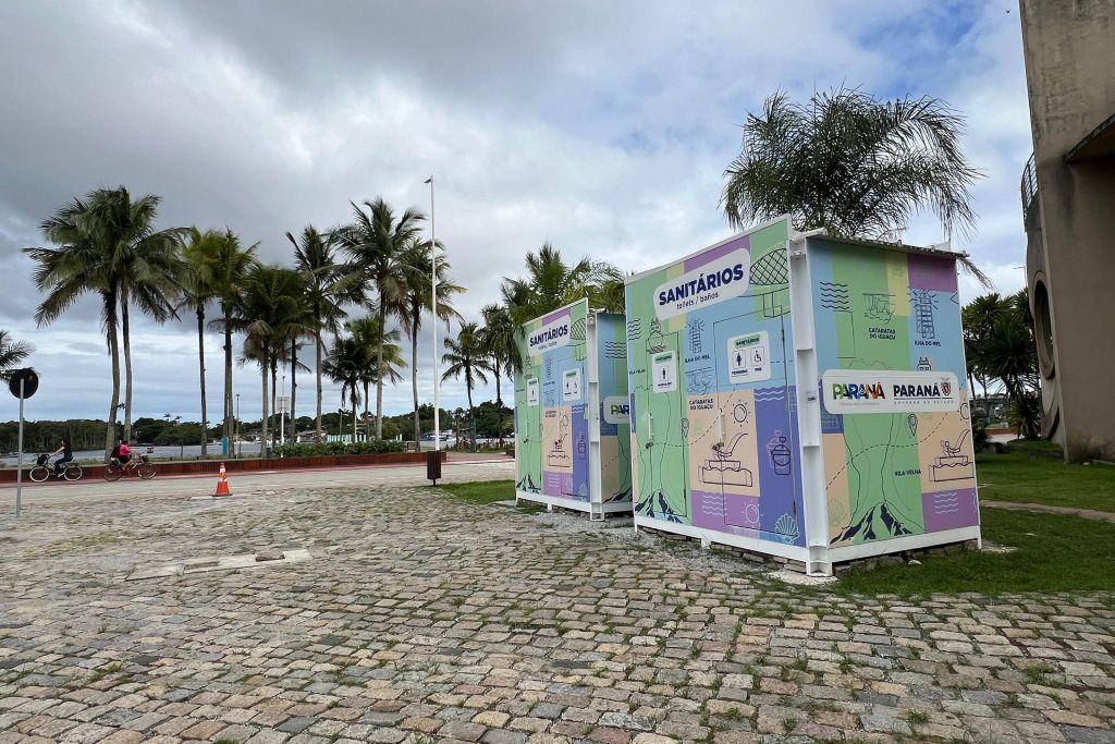 Imagem de sanitários públicos em área aberta, com palmeiras ao fundo e céu nublado. Estruturas coloridas destacam informações sobre serviços e localização.