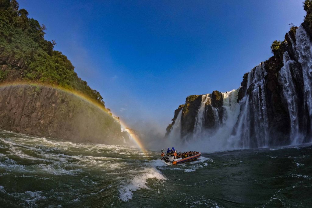 Vista espetacular das Cataratas do Iguaçu com arco-íris ao fundo e um barco em meio à água, ideal para turismo e aventura. Turismo no Paraná cresce quase o triplo da média nacional entre janeiro e outubro.