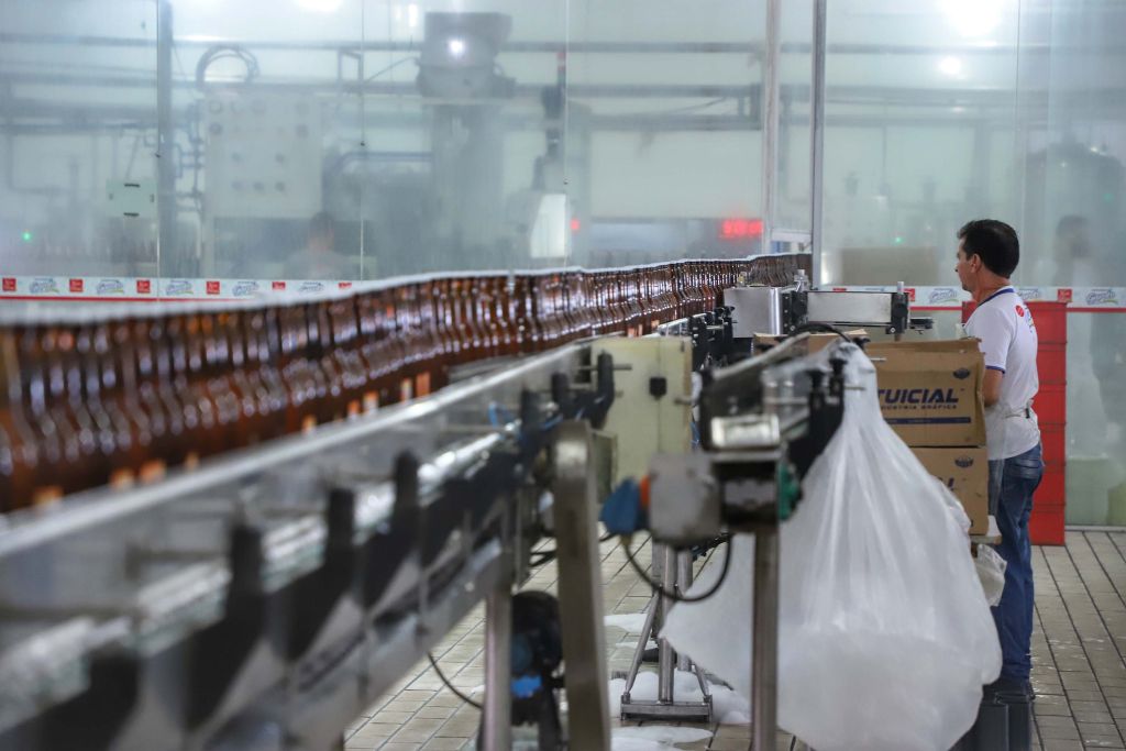 Imagem de uma linha de produção em uma fábrica, com garrafas em movimento e um trabalhador observando o processo. A eficiência na produção é evidente pela organização do ambiente. Indústria do Paraná foi a terceira que mais cresceu no País em outubro, aponta IBGE.
