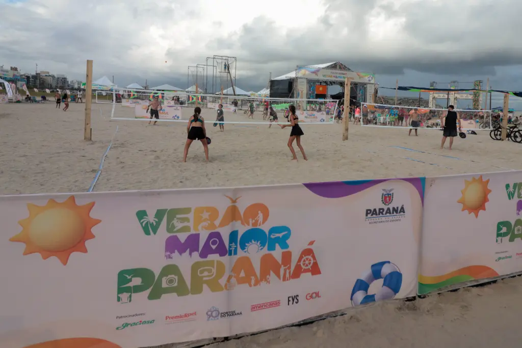 A imagem mostra uma competição de vôlei de praia com várias pessoas jogando em uma areia clara, com uma faixa colorida do evento 'Verão Maior Paraná' ao fundo. Nova infraestrutura das praias paranaenses impressiona turistas estrangeiros
