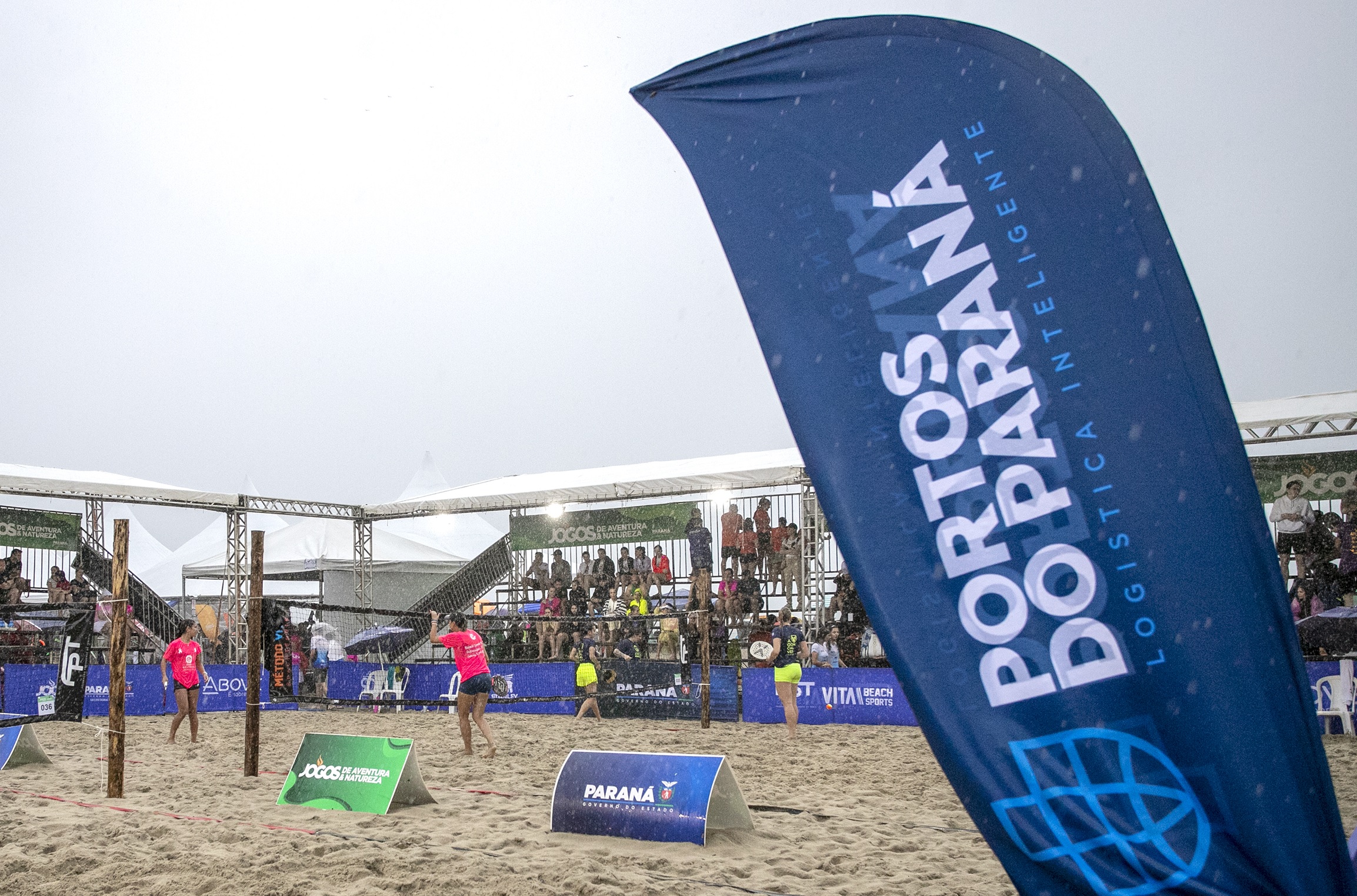 Imagem de um evento de vôlei de praia em Paraná, com destaque para a bandeira 'Portos do Paraná' em um dia chuvoso com público ao fundo.Portos do Paraná traz programação do Verão Maior para o Aeroparque, em Paranaguá