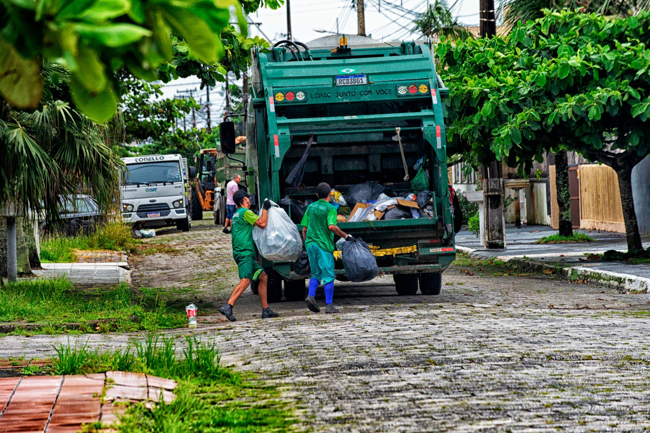 Funcionários da coleta de lixo realizando o trabalho de remoção de resíduos em uma rua suburbanas. A imagem destaca a importância do serviço de limpeza urbana. Com investimento de R$ 15,8 milhões, IAT reforça coleta de resíduos sólidos no litoral