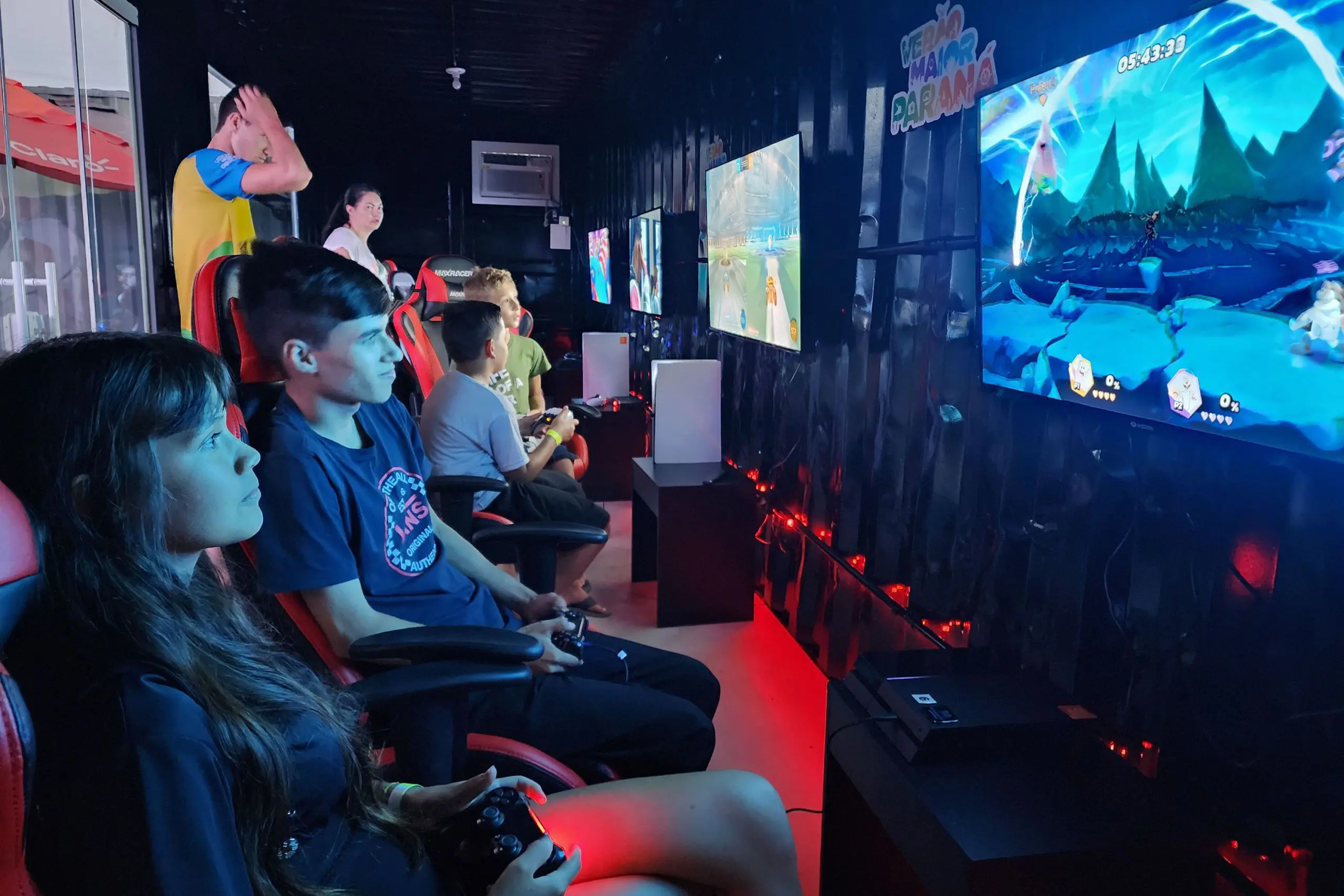 Grupo de jovens jogando videogame em uma sala moderna com várias telas. O ambiente tem iluminação vermelha e cadeiras confortáveis para os jogadores. O foco está no entretenimento digital. Final de torneio de eSports vai agitar Caiobá no domingo no Verão Maior Paraná