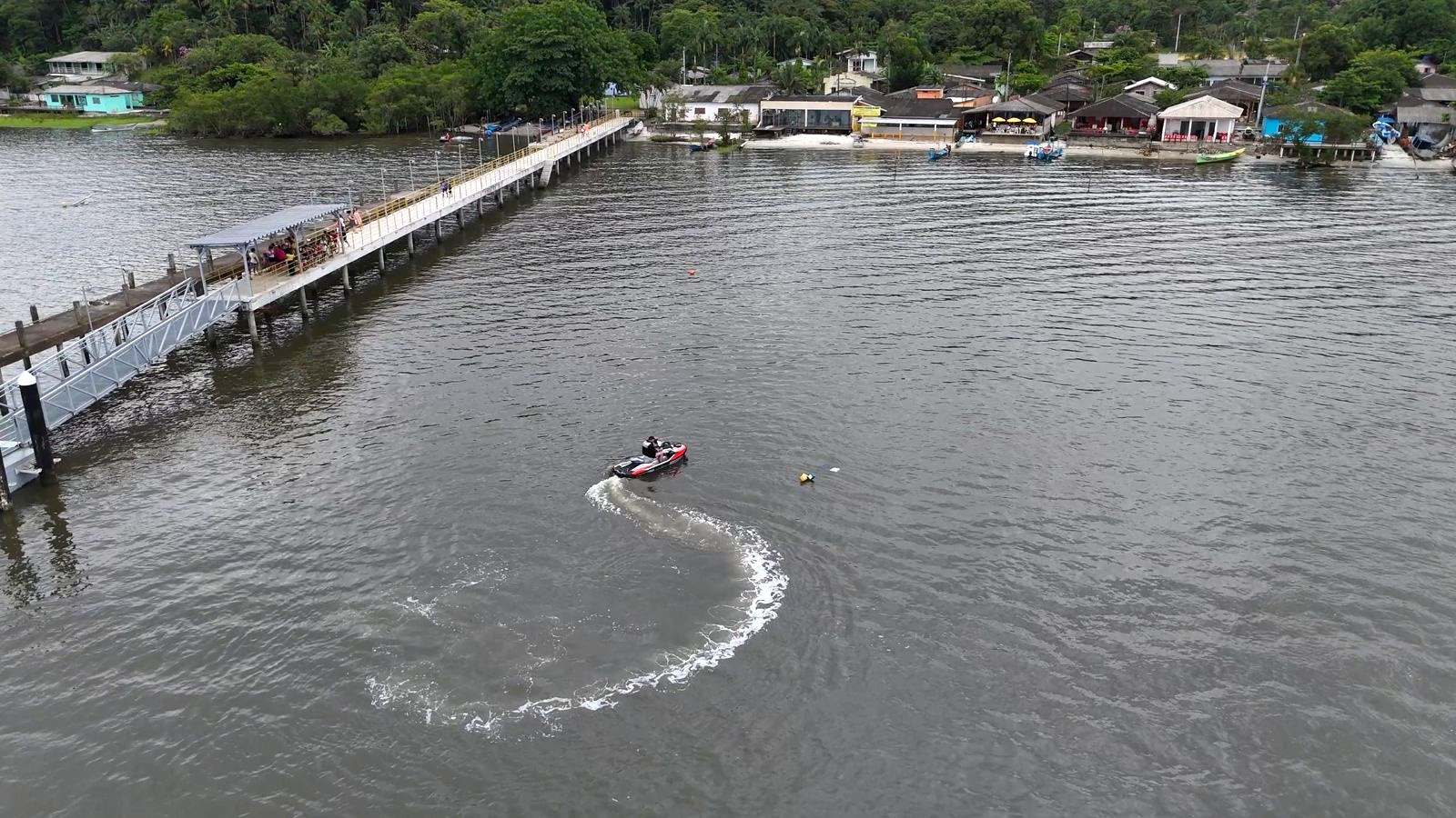 Imagem aérea de uma lancha em um rio, com uma ponte e casas ao fundo, destacando a beleza da natureza e a vida ribeirinha. Festividade de São Sebastião acontece na Ilha do Amparo