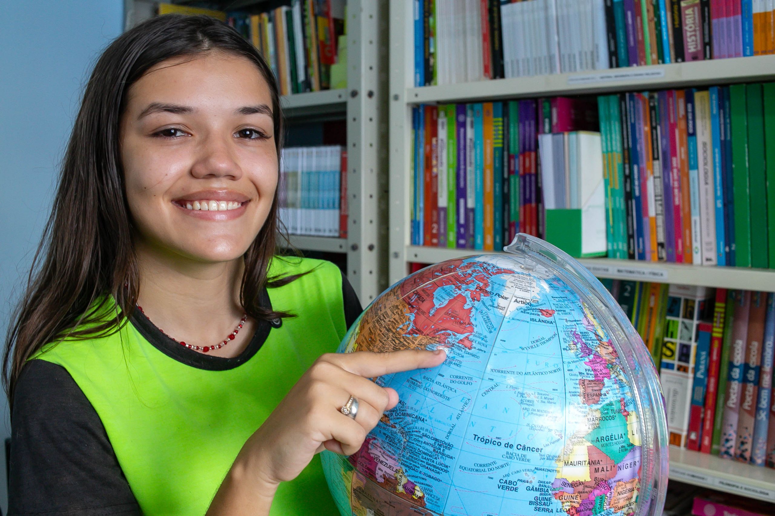 Menina sorridente segurando um globo terrestre em um ambiente de estudo, destacando a geografia e a educação. Da Ilha das Peças para o Canadá: aluna paranaense quer mudar de vida com o Ganhando o Mundo