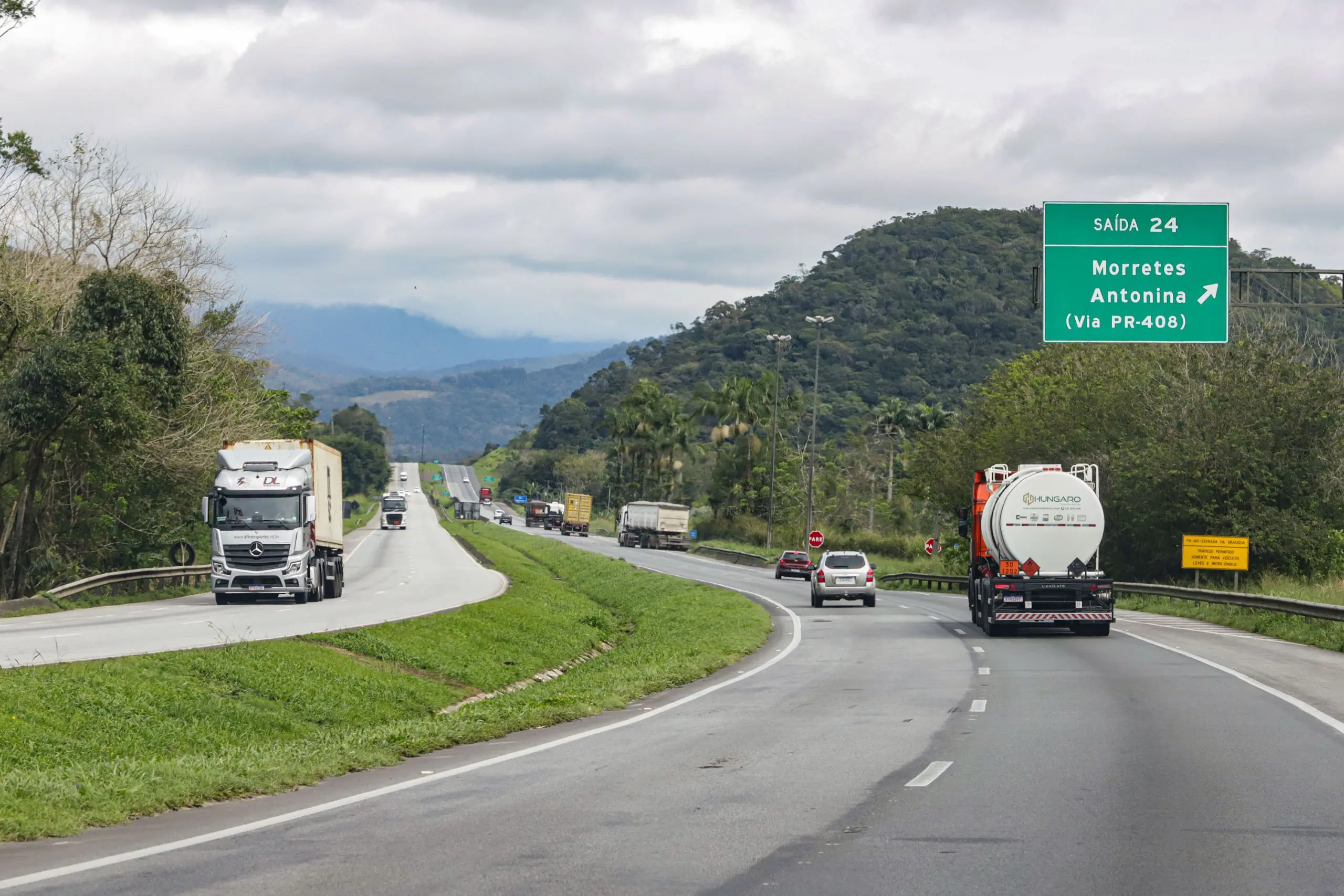 Imagem de uma estrada com caminhões e carros se dirigindo para Morretes e Antonina, PR-408, cercada por montanhas e vegetação. Lote 2: financiamento garante duplicação de 350 km e ampliação da BR-277 na Serra do Mar