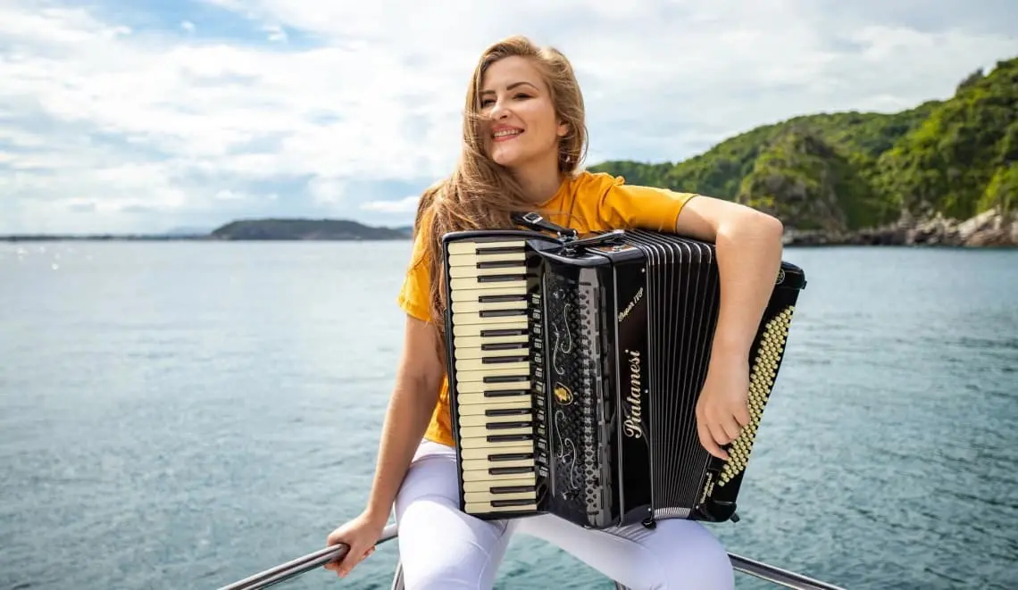 Jovem sorridente vestindo camiseta amarela, tocando acordeão em um barco com vista para o mar e natureza ao fundo em dia ensolarado. Bia Socek tem presença marcada no Palco Sunset em Caiobá, nesta quinta-feira, 16