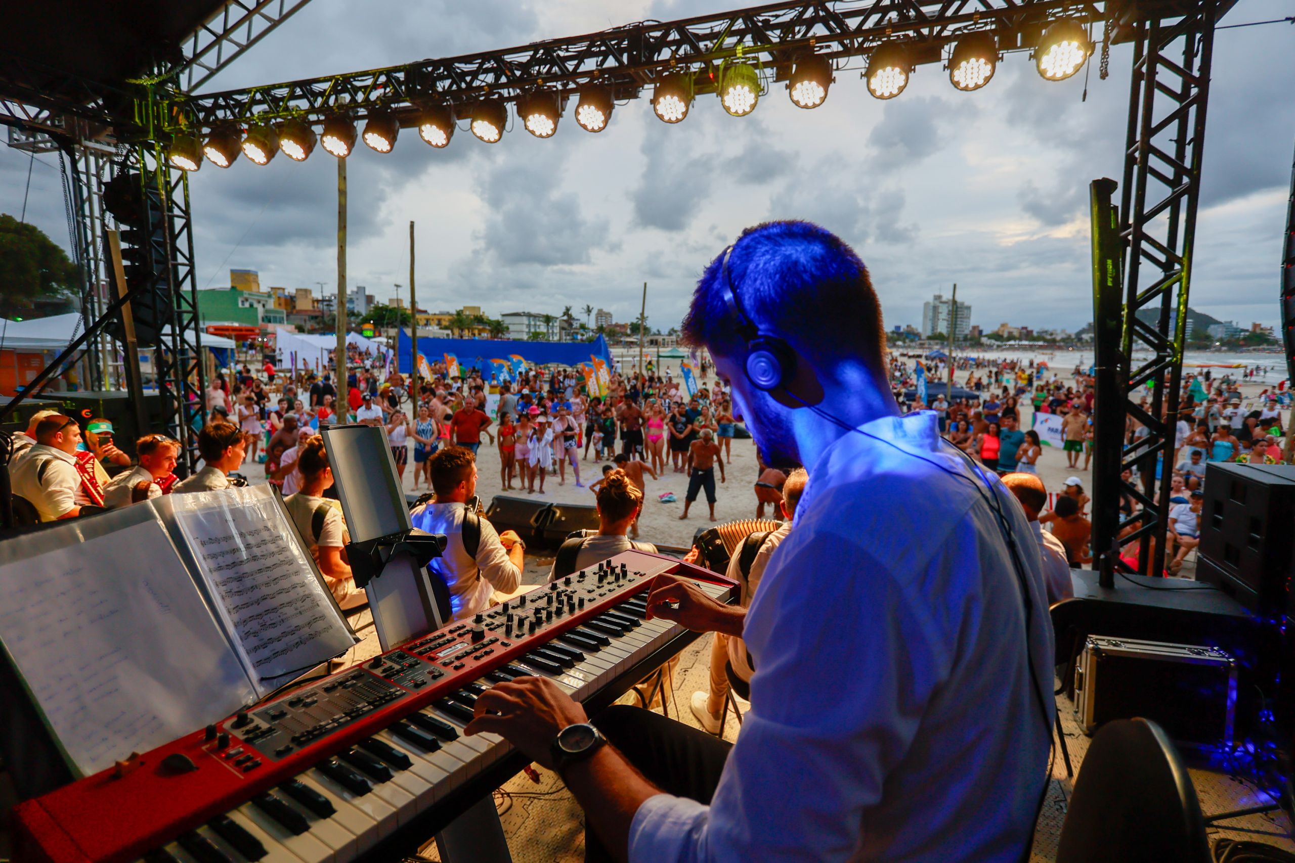 Músico tocando teclado em um palco durante um show na praia, com uma grande multidão se divertindo ao fundo sob um céu nublado. Confira as atrações do Palco Sunset para este fim de semana no litoral do Paraná