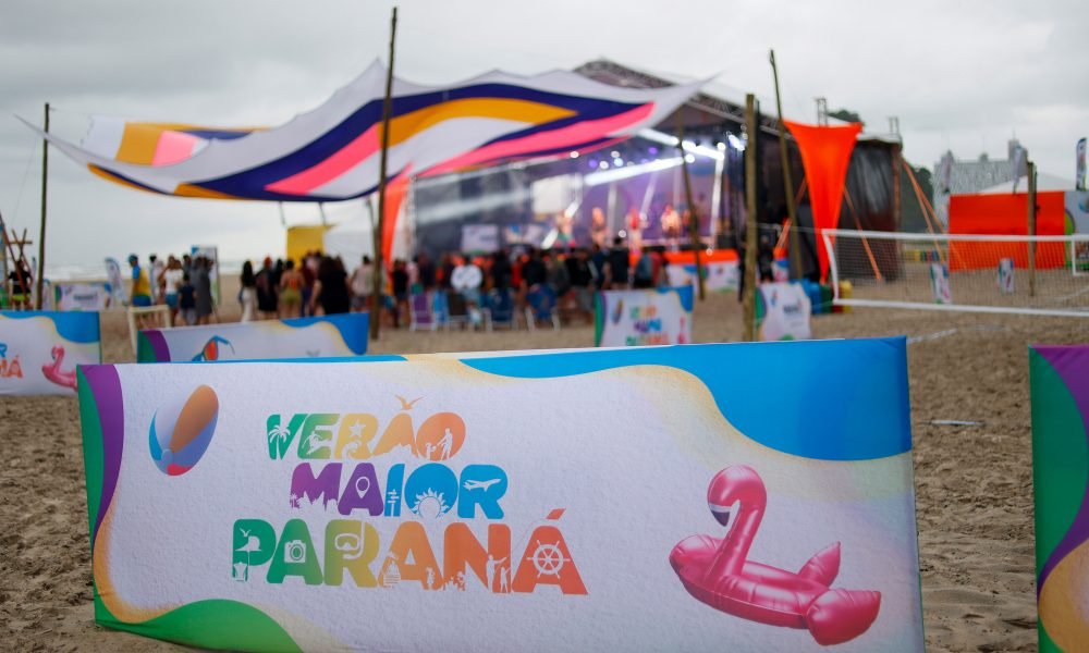 Imagem do evento "Verão Maior Paraná" na praia, com público e estrutura de palco ao fundo. O destaque é para a decoração vibrante e o clima festivo. Banda Milk’n Blues e cantora Poliana levam musicalidade para o Palco Sunset neste sábado