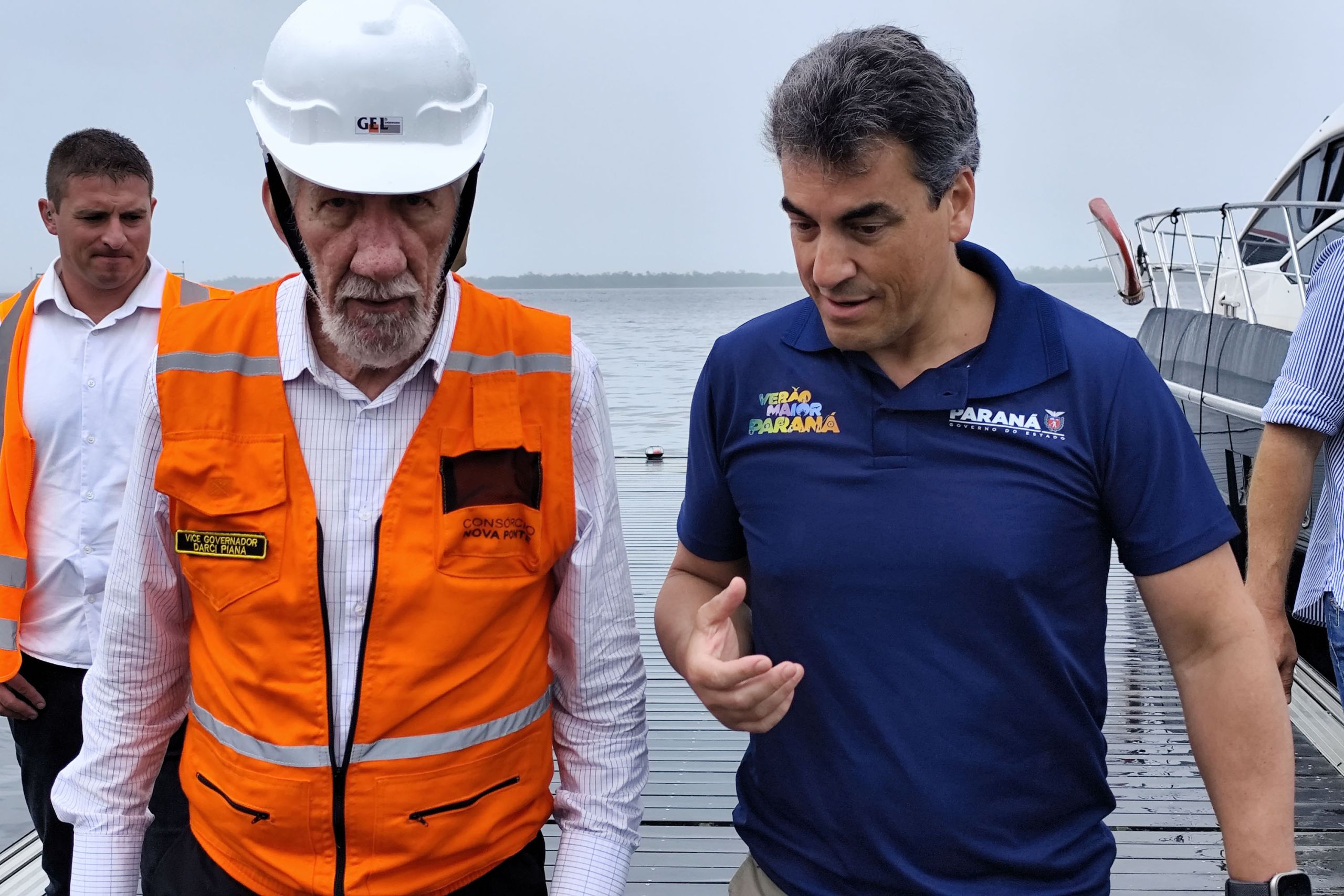 Imagem de dois homens em um local de obras, um deles usando capacete e colete de segurança, discutindo sobre um projeto. O fundo mostra uma área aquática, sugerindo um trabalho de infraestrutura no Paraná. Vice-governador Darci Piana e lideranças acompanham obras da Ponte de Guaratuba