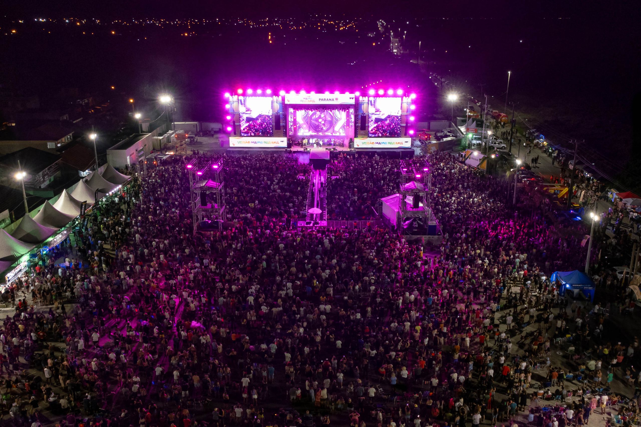 Imagem aérea de um grande show noturno, com um público lotado, palco iluminado em tons de rosa e várias estruturas ao redor, capturando a atmosfera vibrante do evento. Shows do Verão Maior Paraná ultrapassa 800 mil pessoas e encantam artistas nacionais