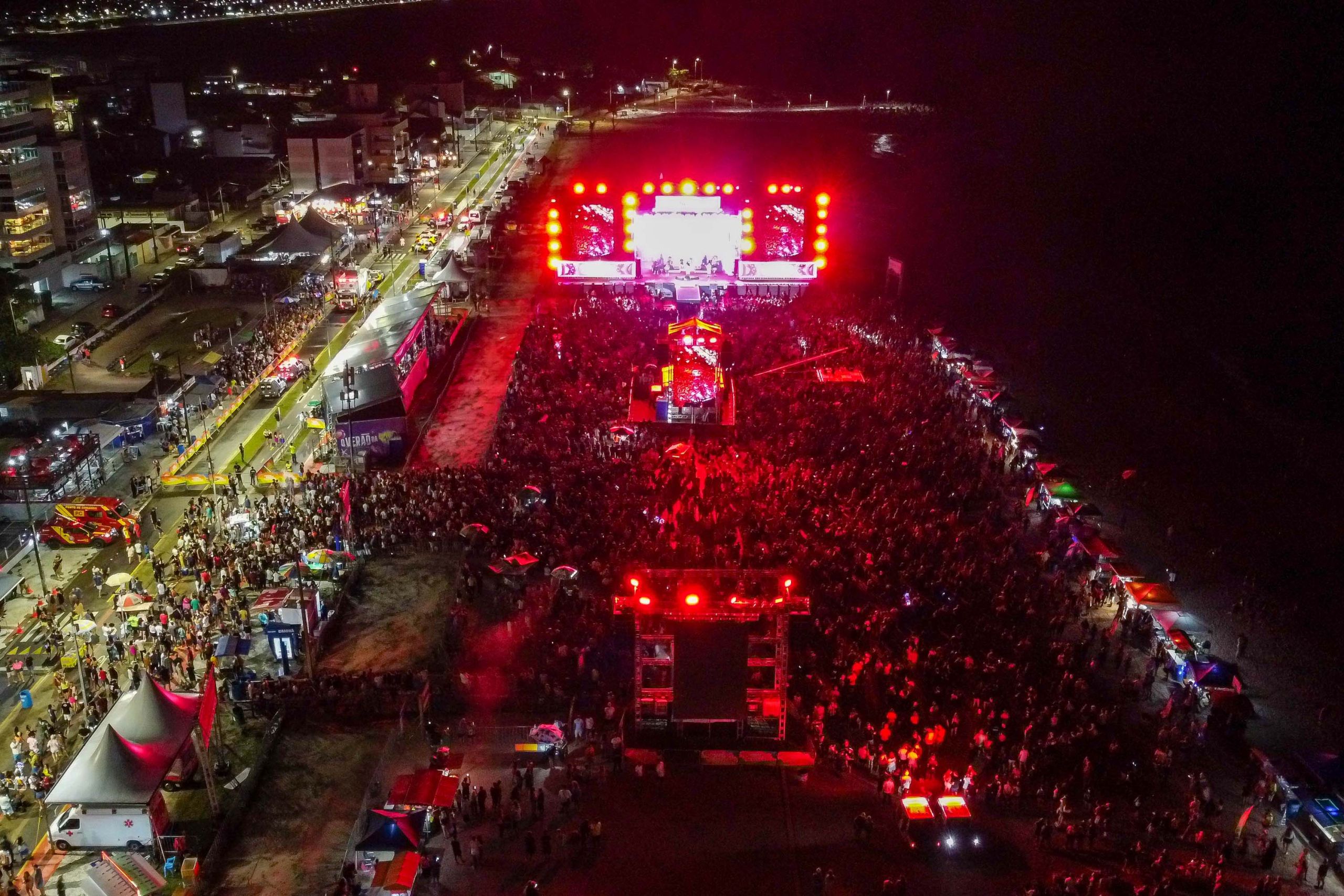 Vista aérea de um evento à noite com multidão e palcos iluminados por luzes vermelhas, destacando a animação e festa no ambiente. Para curtir numa boa: 10 dicas de segurança para ver os shows do Verão Maior Paraná