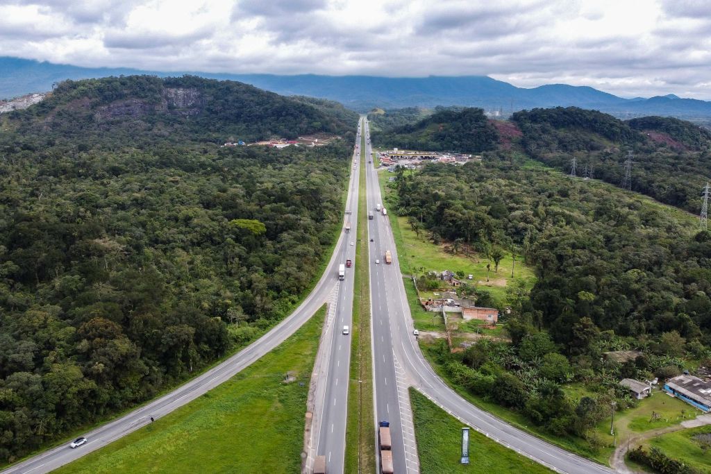 Vista aérea de uma rodovia que se estende entre montanhas e vegetação densa, com tráfego de veículos e nuvens no céu, mostrando a beleza natural da região. Lote 2: financiamento garante duplicação de 350 km e ampliação da BR-277 na Serra do Mar
