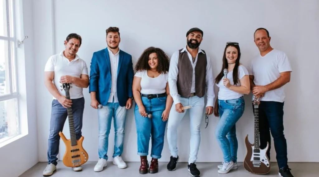 Grupo musical posando em um estúdio com instrumentos. Membros apresentam estilo descontraído e sorridente, transmitindo energia positiva. Cantora Brisan N’Tchalá e a Banda Coração Brasil sobem ao Palco Sunset neste sábado