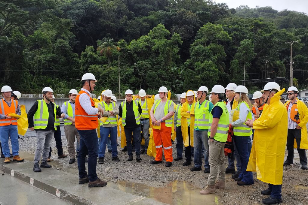 Vice-governador Darci Piana e lideranças acompanham obras da Ponte de Guaratuba 4 Imagem de um grupo de profissionais em um encontro ao ar livre, usando coletes refletivos e capacetes, discutindo sobre segurança no trabalho. Vice-governador Darci Piana e lideranças acompanham obras da Ponte de Guaratuba