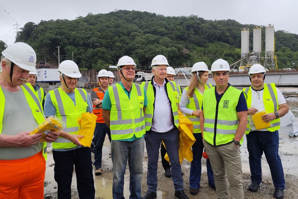 Vice-governador Darci Piana e lideranças acompanham obras da Ponte de Guaratuba 3 Grupo de profissionais em um canteiro de obras, usando coletes de segurança amarelos e capacetes, discutindo projetos em um dia nublado. Vice-governador Darci Piana e lideranças acompanham obras da Ponte de Guaratuba