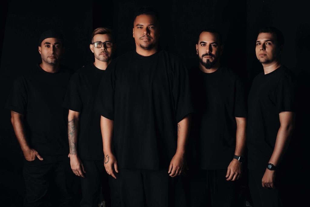 Banda de cinco integrantes vestidos de preto em ambiente escuro, destacando a união e a identidade do grupo. A imagem transmite um clima de seriedade e profissionalismo. Com Luan Santana e Sorriso Maroto, fim de semana terá sertanejo e pagode no litoral
