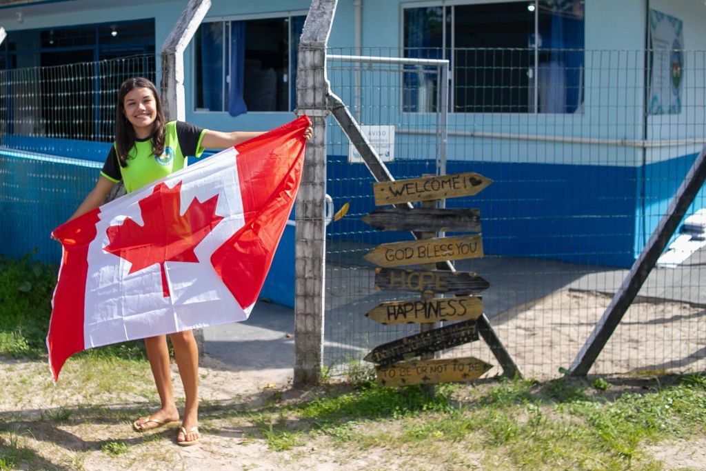 Jovem segurando a bandeira do Canadá em frente a uma estrutura colorida com placa de boas-vindas, simbolizando felicidade e esperança. Da Ilha das Peças para o Canadá: aluna paranaense quer mudar de vida com o Ganhando o Mundo
