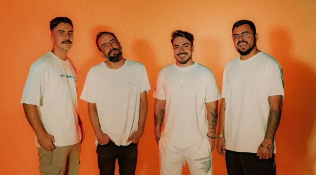 Quatro homens em camisetas brancas posando diante de um fundo laranja, transmitindo uma sensação de amizade e descontração. Eles sorriem e se destacam em um ambiente vibrante. Palco Sunset: Banda Serena Flor e Banda Voraz estreiam nesta terça-feira em Caiobá