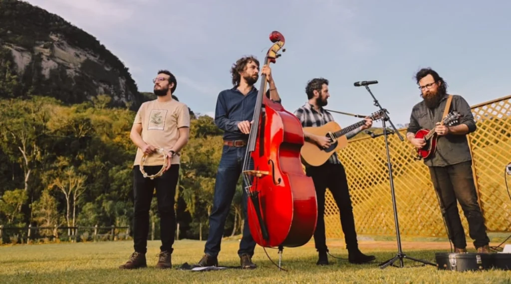 Banda se apresentando ao ar livre, com músicos tocando violão, contrabaixo e guitarra, e um ambiente natural ao fundo. A Banda Lenhadores da Antartida e o cantor Peci agitam o Palco Sunset nesta sexta-feira
