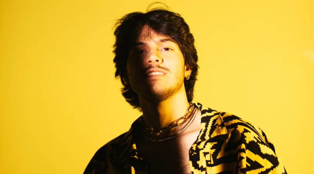 Imagem de um jovem com cabelo ondulado, vestindo uma camisa estampada em preto e amarelo, posando em frente a um fundo amarelo vibrante. A Banda Lenhadores da Antartida e o cantor Peci agitam o Palco Sunset nesta sexta-feira
