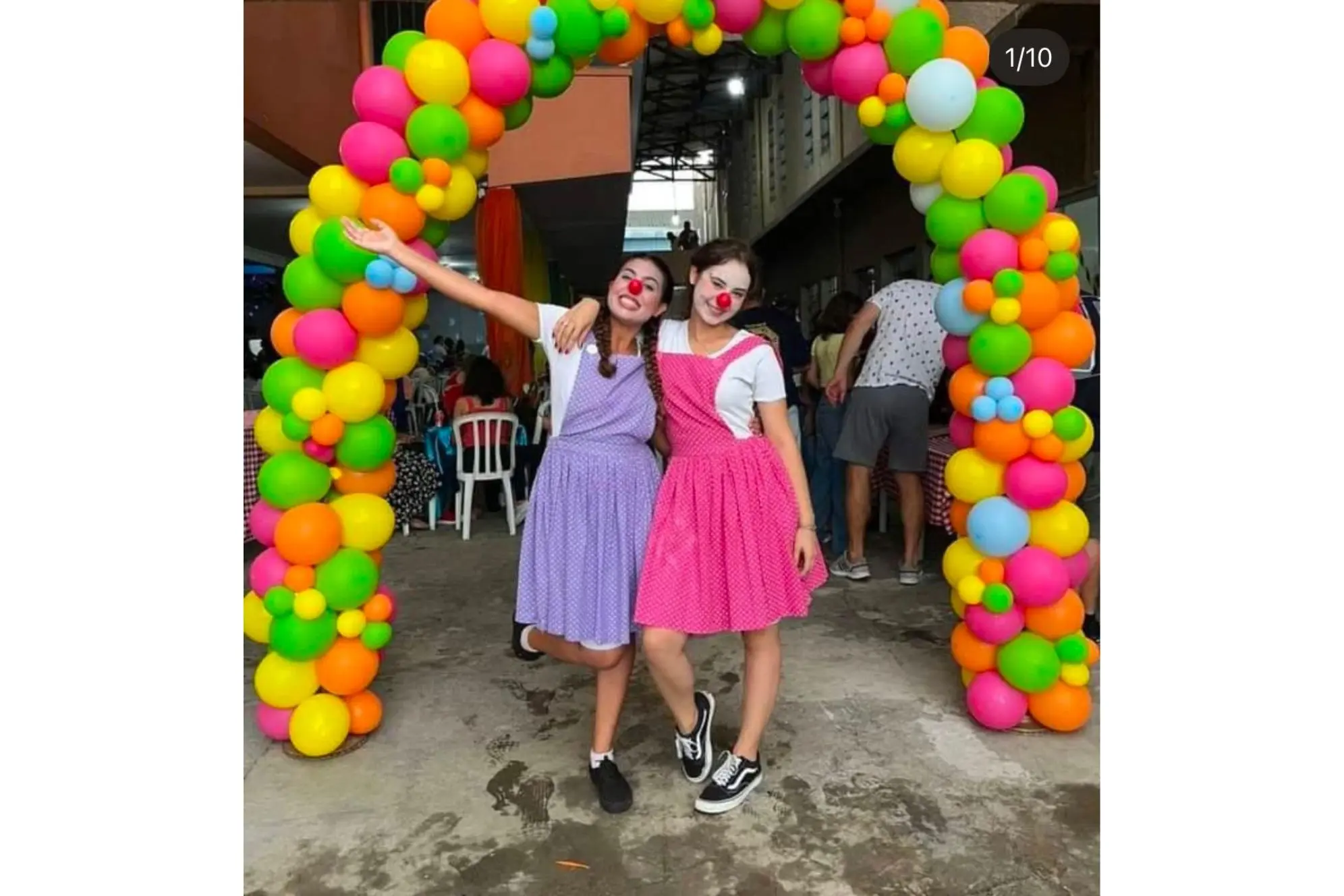 Duas meninas vestidas como palhaças, sorrindo e posando sob um arco de balões coloridos em uma festa animada. Renovação Carismática Católica promove o Cristoval 2025 no dia 4 de março