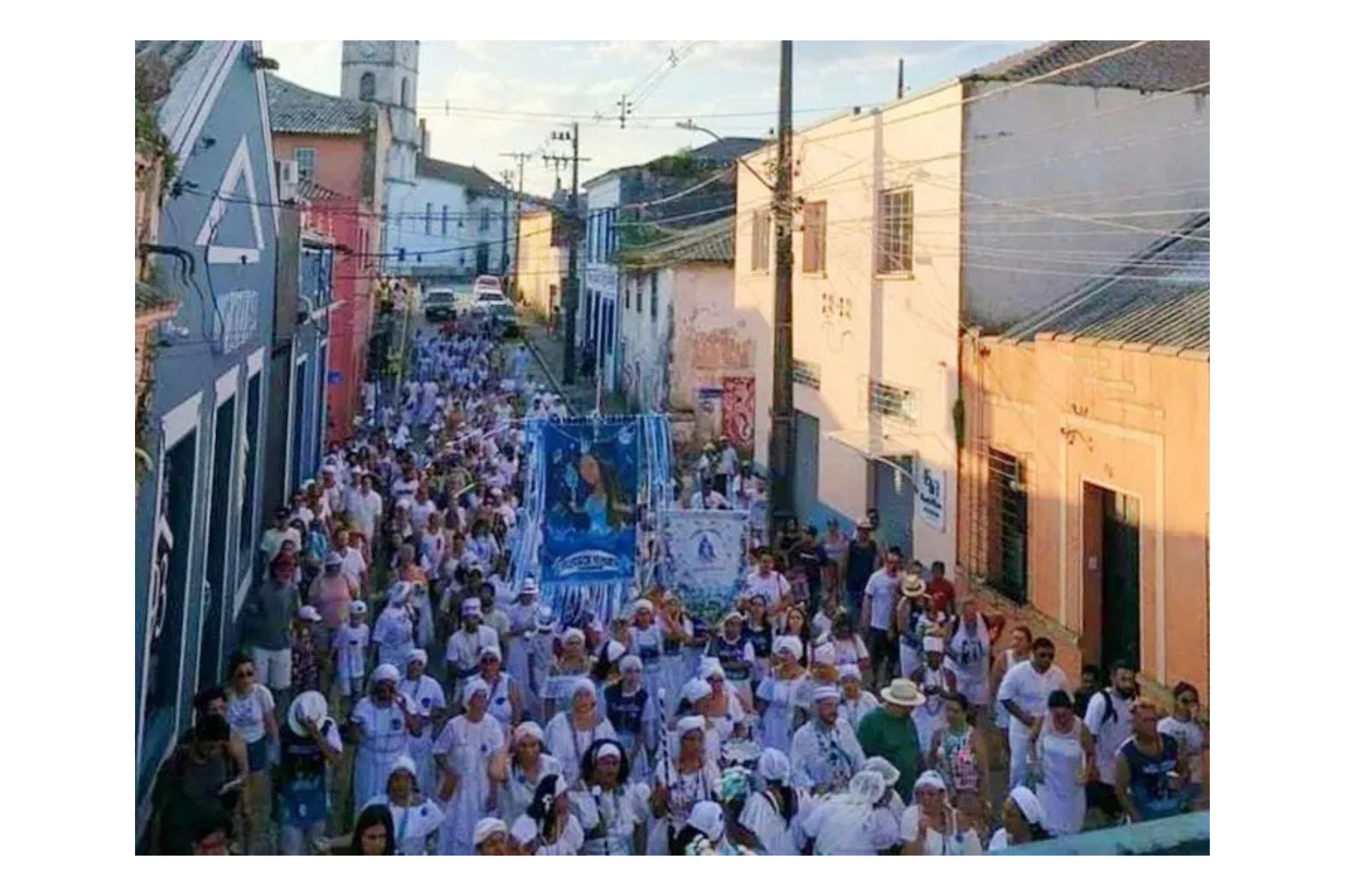 Uma grande multidão de pessoas vestidas de branco participa de um desfile em uma rua, segurando bandeiras e objetos religiosos, simbolizando uma festa tradicional. 7.º Afoxé Filhos de Iemanjá acontecerá em Paranaguá