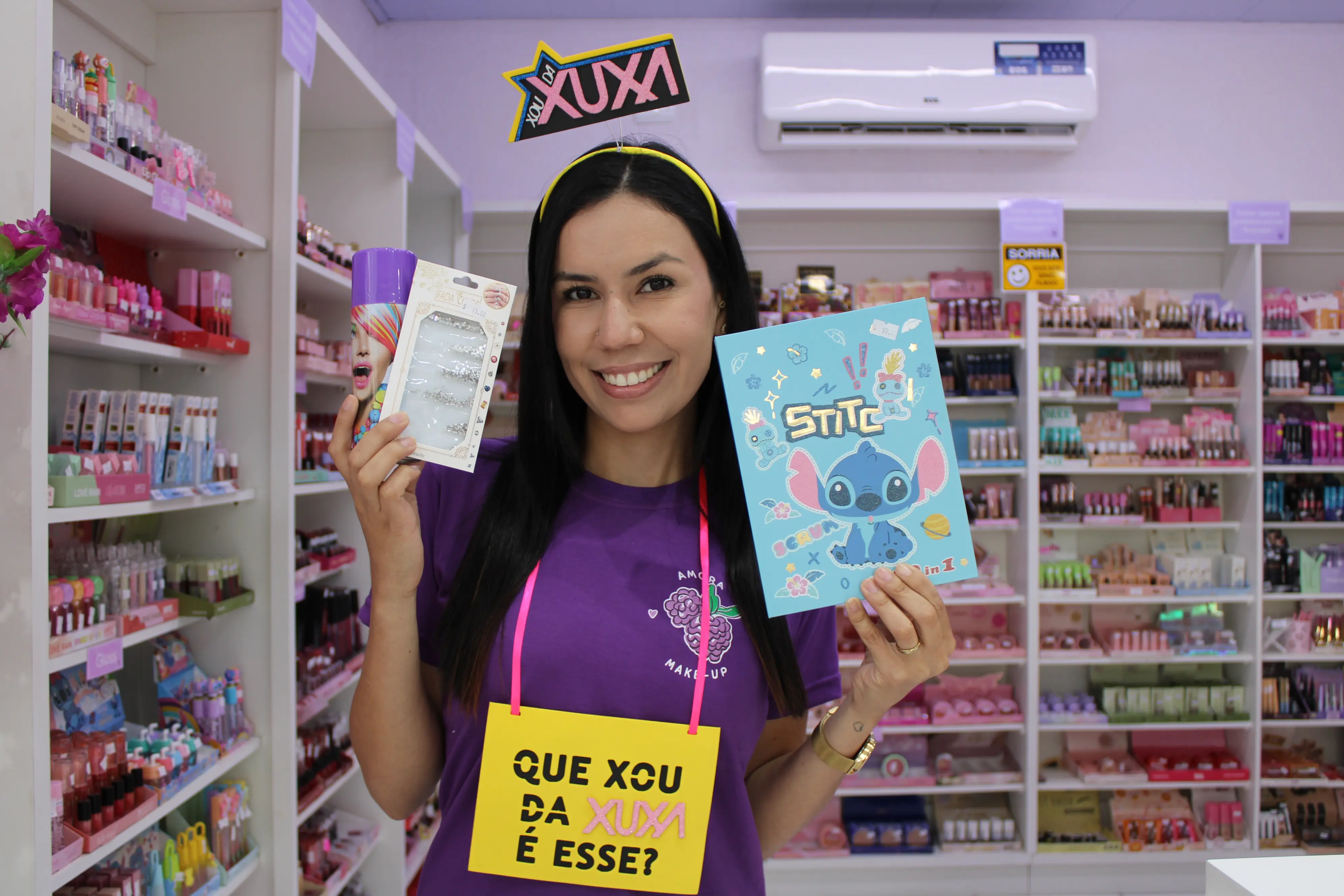 Mulher sorridente segurando produtos de beleza e um caderno com Stitch em uma loja de cosméticos, com destaque para o tema da Xuxa. Confira dicas para brilhar com abadás e acessórios no Carnaval