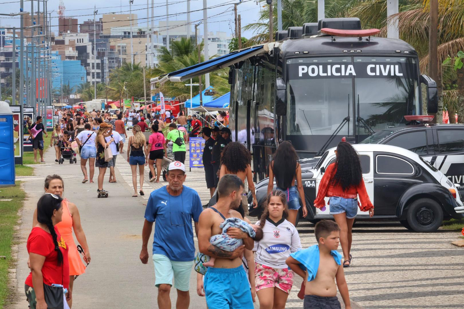 Avenida movimentada com muitas pessoas durante um evento, com policiamento visível e atividades ao ar livre, destacando a presença da Polícia Civil. Delegacia Móvel da PCPR é ponto de referência nos eventos do Verão Maior Paraná