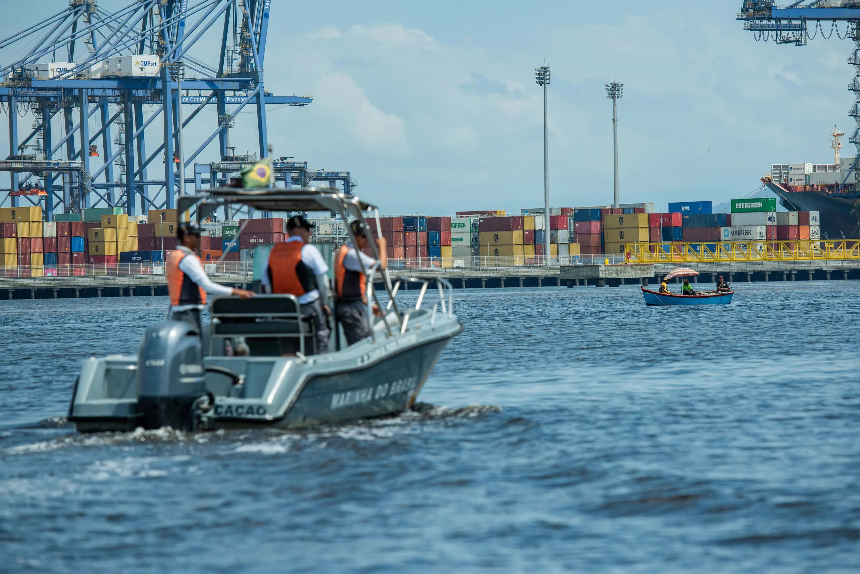 Imagem de uma embarcação em movimento no porto, com containers e guindastes ao fundo, representando a atividade no setor portuário. Fiscalização marítima é realizada nos portos paranaenses